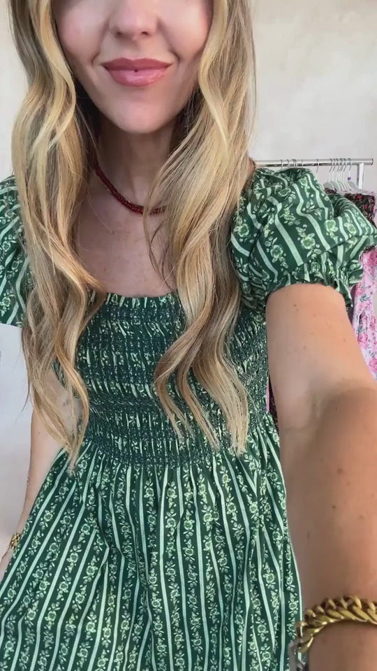 A woman with long, wavy blonde hair wears the Ivy City Co Sunday Best Dress in Pine Felicity Floral—featuring white floral stripes—paired with a red necklace and a gold bracelet. Designed for nursing and maternity, she smiles as she takes an indoor selfie, clothing racks visible behind her.