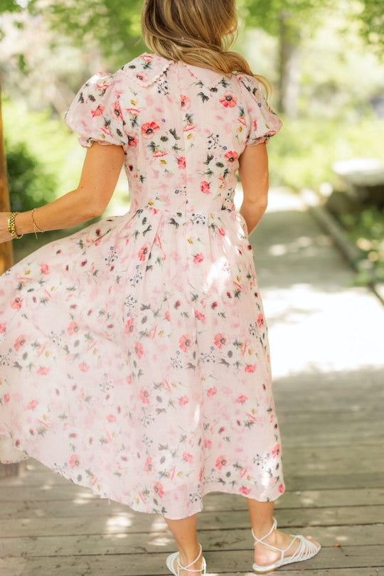 A woman stands outdoors, facing away, wearing the Ivy City Co Sophia Dress in Roselyn Pink—a light pink, floral-print cotton-linen midi dress with puff sleeves—paired with white sandals. She holds part of her dress as she stands on a wooden path surrounded by greenery.