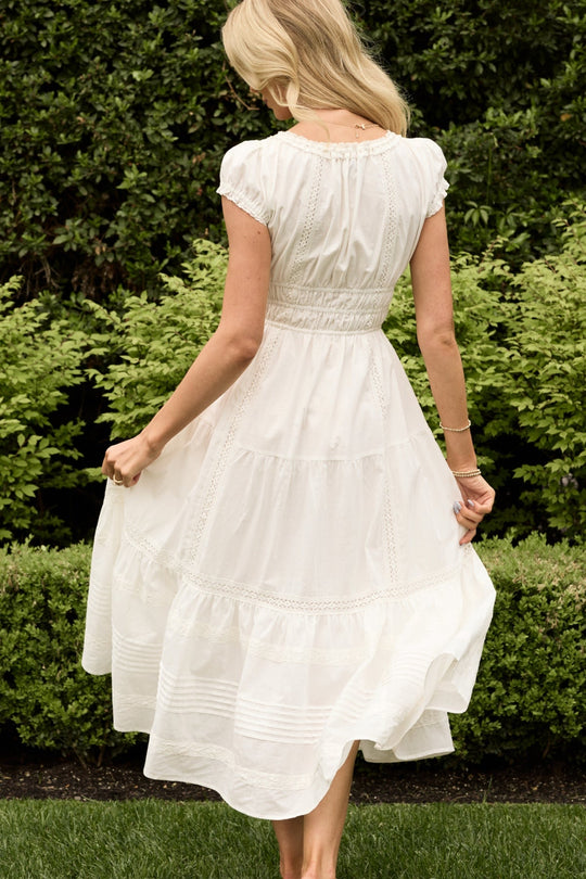 A woman stands barefoot on the grass, facing away and holding her skirt, wearing the Ivy City Co Palermo Dress in White—a flowy, nursing-friendly style. Lush green bushes and trees fill the background.