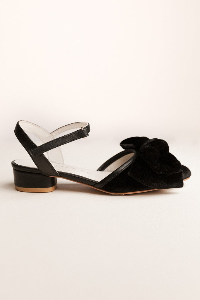 Showcased against a plain light background, the Mini Eloise Bow Heel in Black by Ivy City Co features a low heel, ankle strap, and a statement bow detail on the front for an elegant look.