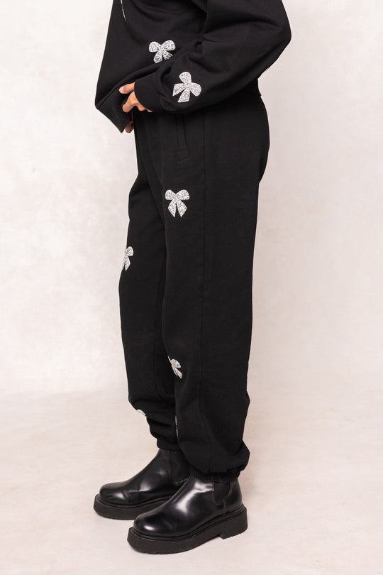 A person is shown from the shoulders down, wearing the Ivy City Co Micah Jogger Sweatpants in Black, which feature sparkling rhinestone bow designs. The joggers are styled with black boots and a matching black top, with the individual standing on a light floor against a neutral background.