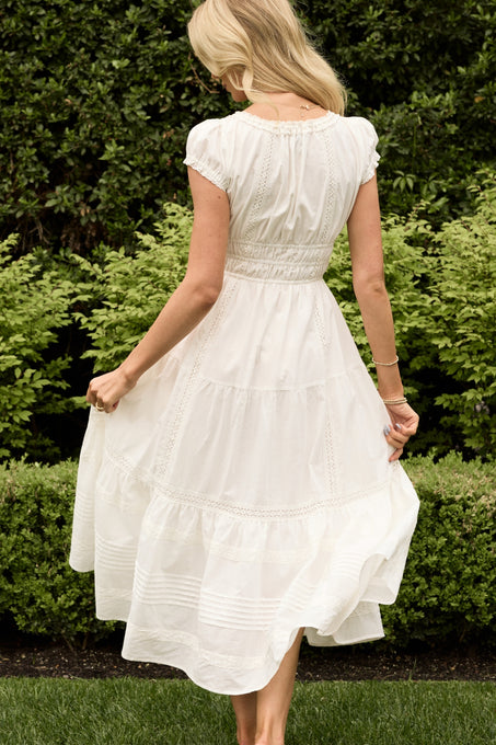 A woman wears the Ivy City Co Palermo Dress in White, standing barefoot on grass and gently twirling, surrounded by lush green bushes and trees in a garden.