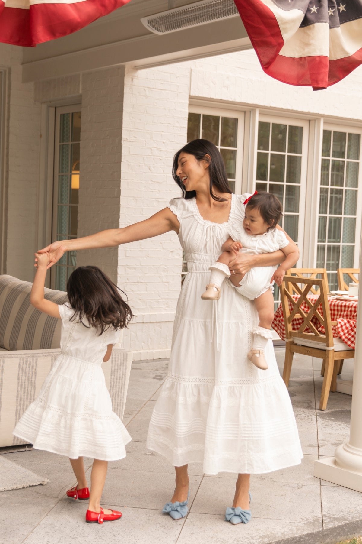A woman in the Ivy City Co Palermo Dress in White twirls a young girl in a matching outfit while holding a baby, all dressed in white, on a patio with red, white, and blue bunting—maternity-friendly style perfect for family moments.