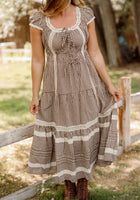 A woman stands outside by a wooden fence, holding her skirt while wearing the Ivy City Co Palermo Dress in Hickory Gingham, featuring lace trim and charming puff sleeves. She pairs the maternity-friendly dress with brown lace-up boots, and her face is not visible.