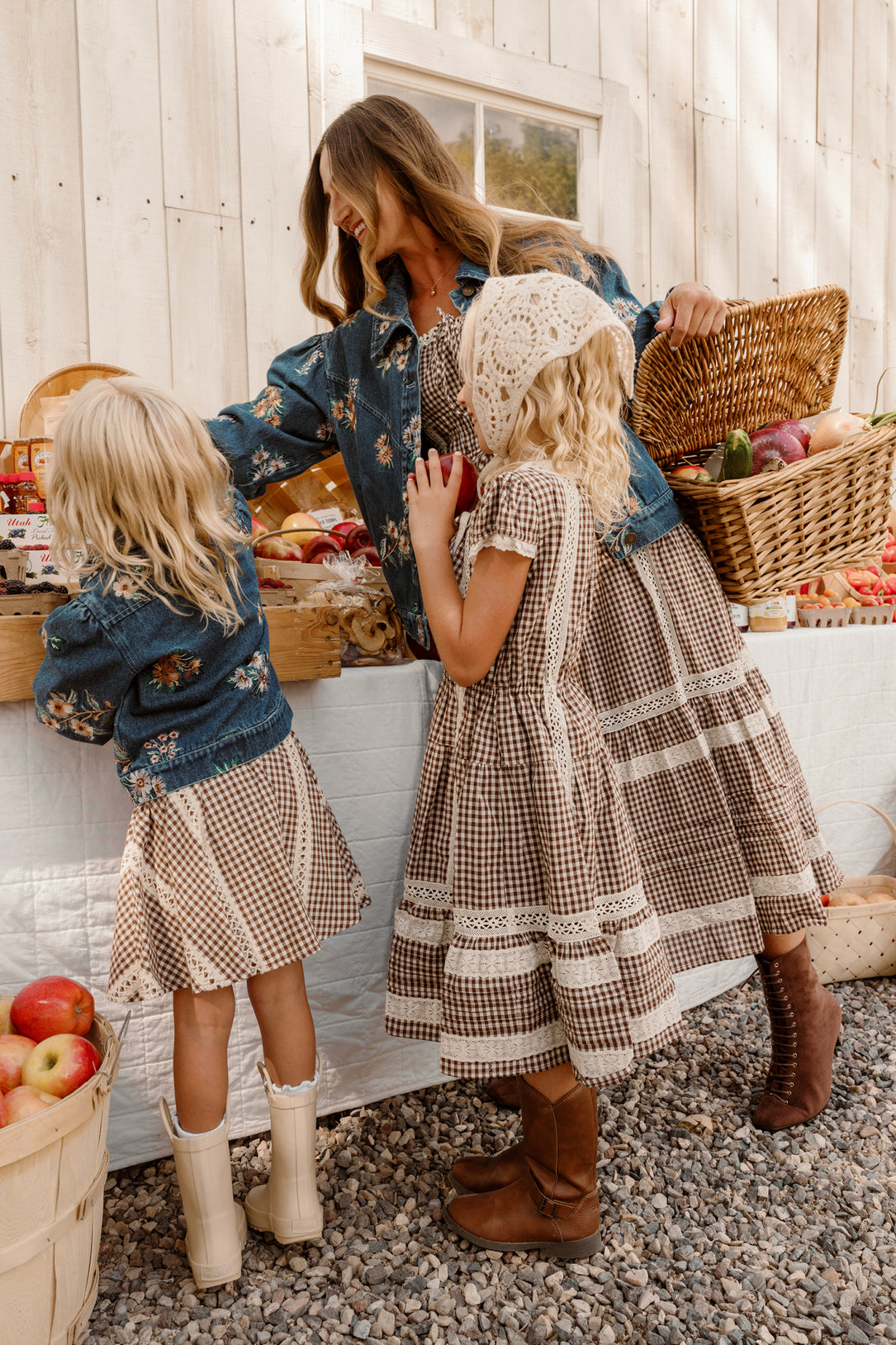 A woman and two young girls wear matching Ivy City Co Palermo Dresses in Hickory Gingham with puff sleeves as they shop for produce at an outdoor market stand, surrounded by baskets of vegetables and apples—their coordinated outfits are both stylish and maternity friendly.