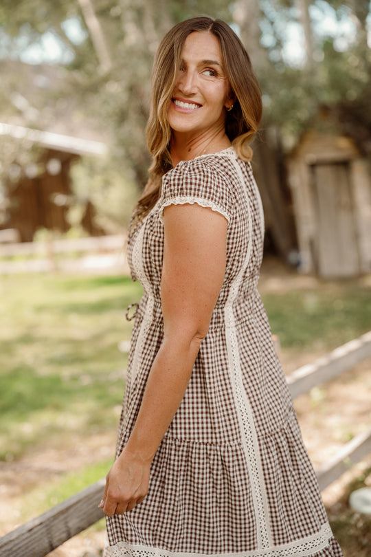A woman with long brown hair poses outdoors, smiling at the camera in the Ivy City Co Palermo Dress in Hickory Gingham. This maternity-friendly look features puff sleeves and is set against a backdrop of trees, grass, and a rustic wooden structure blurred in the background.