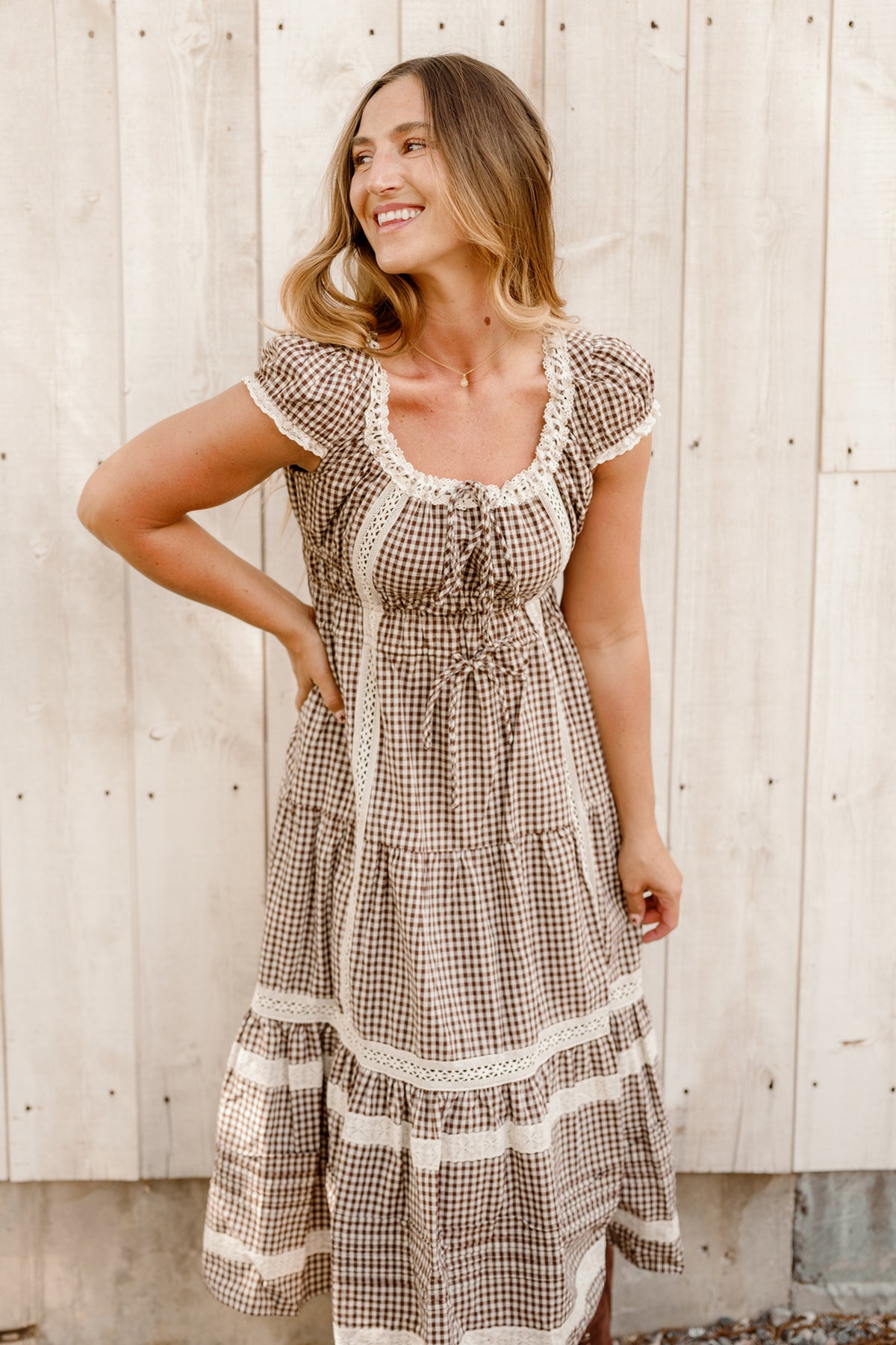 A woman stands smiling in front of a light wooden wall, wearing the Ivy City Co Palermo Dress in Hickory Gingham—a brown and white checkered style with lace details, short puff sleeves, and a maternity-friendly fit.