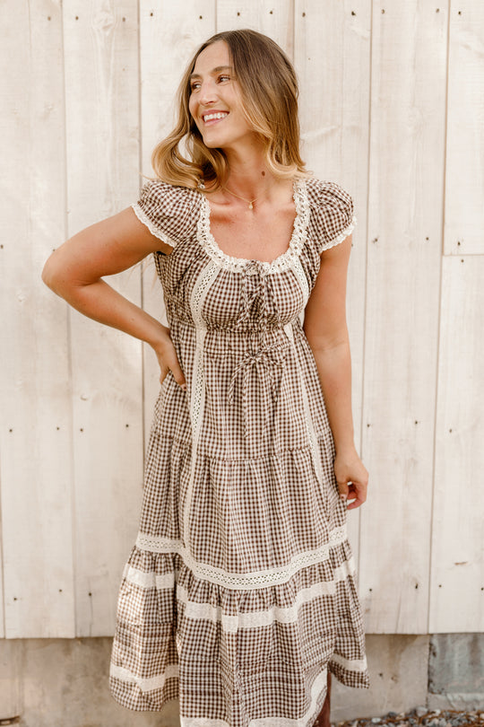 A woman stands smiling in front of a light wooden wall, wearing the Ivy City Co Palermo Dress in Hickory Gingham—a brown and white checkered style with lace details, short puff sleeves, and a maternity-friendly fit.