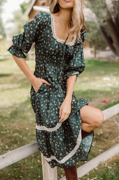 Wearing the Palermo 3/4 Sleeves Dress in Evergreen Lovelorn Ditsy by Ivy City Co, a woman poses outdoors by a wooden fence, with one knee raised and her hand in her pocket. The maternity-friendly cotton dress, adorned with floral details and lace trim, complements the natural setting of trees and a distant house softly blurred in the background.