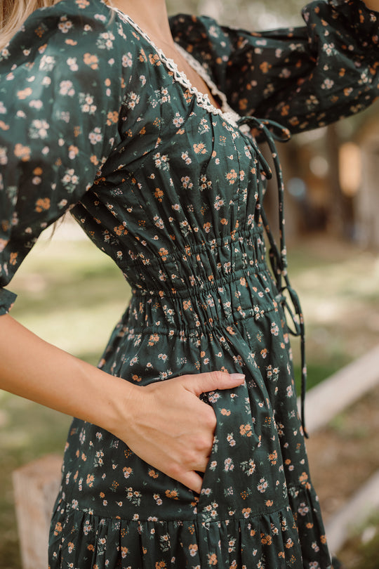 Wearing the Palermo 3/4 Sleeves Dress in Evergreen Lovelorn Ditsy by Ivy City Co, a woman stands outdoors with one hand in her pocket, highlighting the dress’s ruched waist and delicate ditsy floral pattern. With puffed sleeves and lace trim, this maternity-friendly style blends comfort and elegance for any occasion.