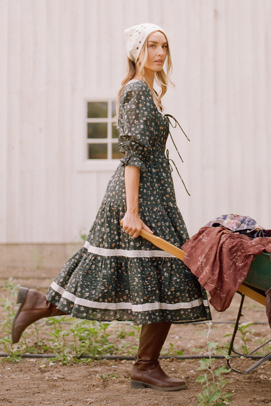 A woman wearing the Ivy City Co Palermo 3/4 Sleeves Dress in Evergreen Lovelorn Ditsy, paired with brown boots and a white headscarf, pushes a green wheelbarrow filled with clothes in front of a white building with a window.