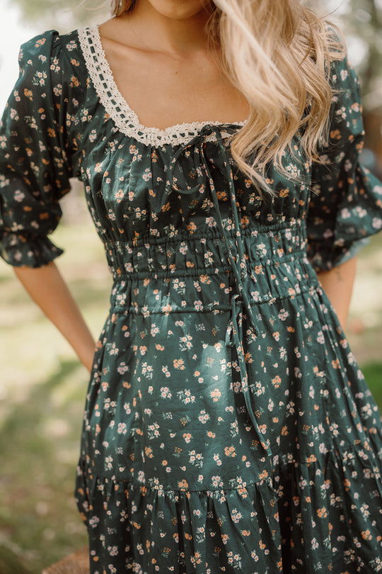 A woman stands outdoors in the Palermo 3/4 Sleeves Dress in Evergreen Lovelorn Ditsy by Ivy City Co, featuring a dark green floral pattern and a lace-trimmed square neckline. Her long, wavy blonde hair cascades over her shoulder, and the dress’s comfortable fit makes it maternity friendly.