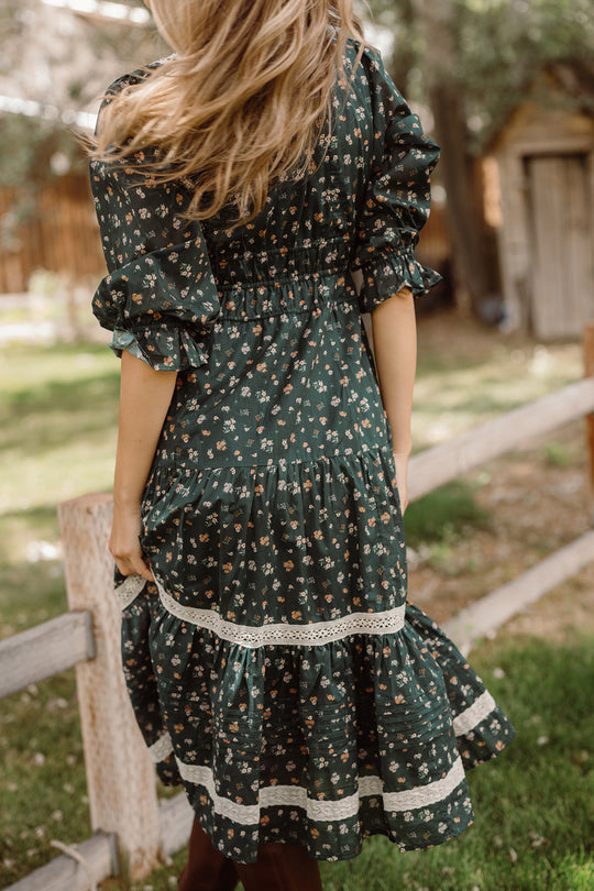 A woman with long hair is standing outdoors by a wooden fence, holding the skirt of her Ivy City Co Palermo 3/4 Sleeves Dress in Evergreen Lovelorn Ditsy. The floral cotton dress features lace trim and 3/4 sleeves, creating a graceful and maternity-friendly look amid trees and a wooden shed in the background.