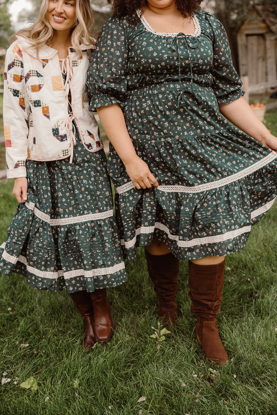 Two women stand on the grass, both smiling warmly in matching Ivy City Co Palermo 3/4 Sleeves Dresses in Evergreen Lovelorn Ditsy, featuring floral cotton fabric, lace trim, and a maternity-friendly design. One layers her dress with a patchwork jacket, while the other completes her look with brown knee-high boots. Trees and a wooden building set a sunny outdoor backdrop for their coordinated outfits.