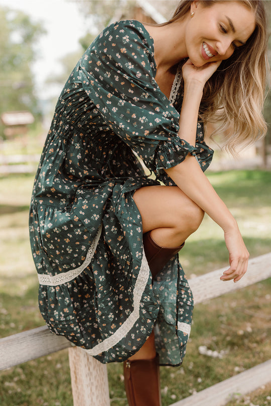 A woman wearing the Palermo 3/4 Sleeves Dress in Evergreen Lovelorn Ditsy by Ivy City Co, paired with brown boots, sits on a wooden fence outdoors. She smiles and looks down, surrounded by green trees and grass. The stylish dress is also maternity friendly.