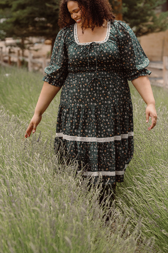 Dressed in the Palermo 3/4 Sleeves Dress in Evergreen Lovelorn Ditsy by Ivy City Co, a woman strolls gracefully through a field of tall grass or lavender, her hand gently brushing the plants. The maternity-friendly silhouette enhances her elegance, with trees and a fence visible in the background.