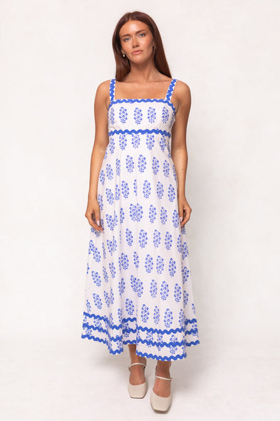 A woman with long brown hair and white shoes models the Ivy City Co Palmer Dress in Blue Block Print, a chic, sleeveless, ankle-length cotton midi dress with decorative blue trim. The design is stylish and maternity friendly.