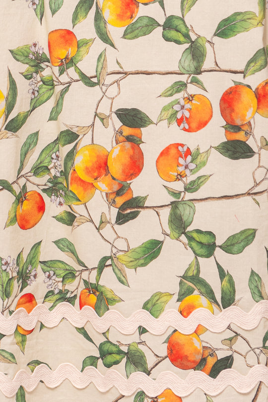 The Palmer Dress in Peach Blossom by Ivy City Co features illustrated oranges, green leaves, white blossoms, and wavy cream trim—perfect for a cotton midi dress that’s also maternity friendly.