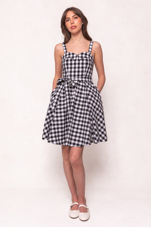 A woman with long, wavy brown hair stands against a light background, wearing the Ivy City Co Patty Dress in Black Gingham with a waist bow and white flats. She has her hands in the dress’s pockets.