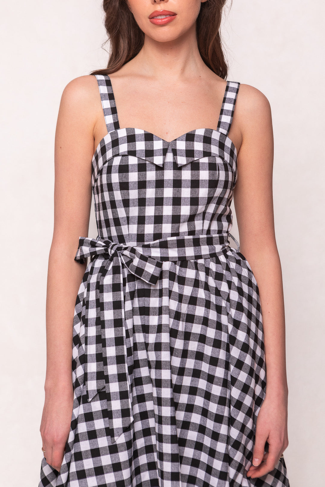 A woman wears the Ivy City Co Patty Dress in Black Gingham, featuring a sweetheart neckline and matching waist tie, standing against a plain background.