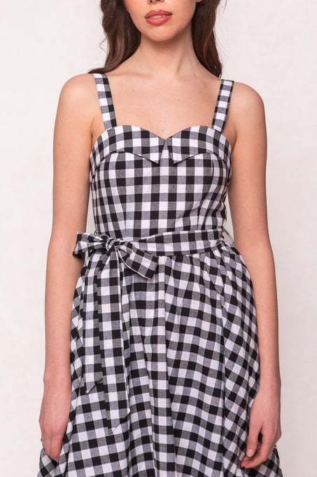 A woman wears the Ivy City Co Patty Dress in Black Gingham, featuring a sweetheart neckline and matching waist tie, standing against a plain background.