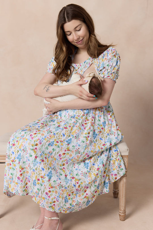 Wearing the Ivy City Co Amelie Dress in Floral Fields, a woman sits on a bench, gently cradling and gazing down at a swaddled newborn baby in her arms. The background is neutral and soft.