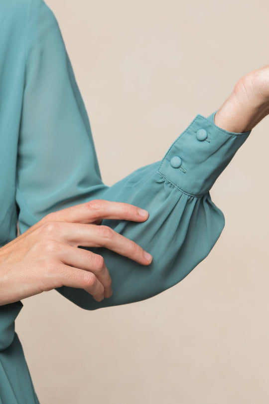 A person wearing the Andie Dress in Seaglass Chiffon by Ivy City Co gently touches their sleeve, highlighting the fabric-covered cuff buttons against a neutral background, evoking an elegant and refined style.