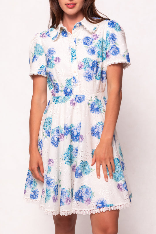 A woman wears the Ivy City Co Penny Dress in Blue Floral, a collared knee-length dress featuring blue and purple florals, button-down front, and lace-trimmed sleeves and hem—blending elegance with charm.