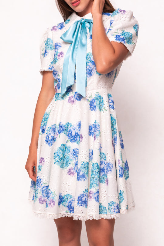 A woman models the Ivy City Co Penny Dress in Blue Floral, featuring cotton eyelet fabric, short puff sleeves, lace trim, and a large light blue collar bow. She poses against a plain background with one hand touching her hair.