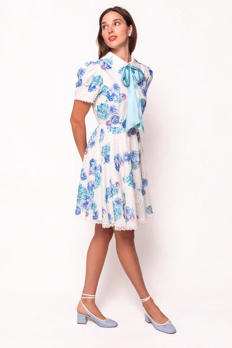 A woman models the Ivy City Co Penny Dress in Blue Floral, featuring a large blue bow at the collar and paired with light blue heels. She stands against a white background, looking slightly to the side with her hands behind her back.