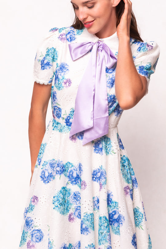 A woman wears the Penny Dress in Blue Floral by Ivy City Co, featuring cotton eyelet fabric, blue and purple floral prints, short puff sleeves, and a large lavender bow at the collar as she stands against a light background, smiling.