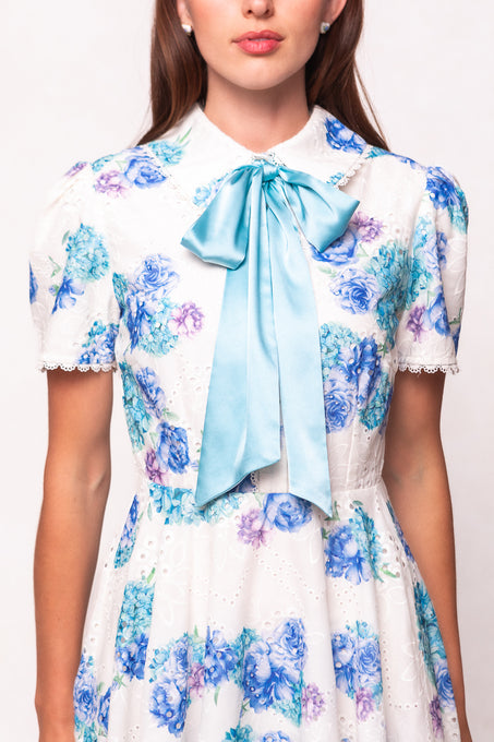 A woman wears the Ivy City Co Penny Dress in Blue Floral, a short-sleeve cotton eyelet dress with lace trim and a large light blue satin bow at the collar. The image is cropped at her shoulders and knees.