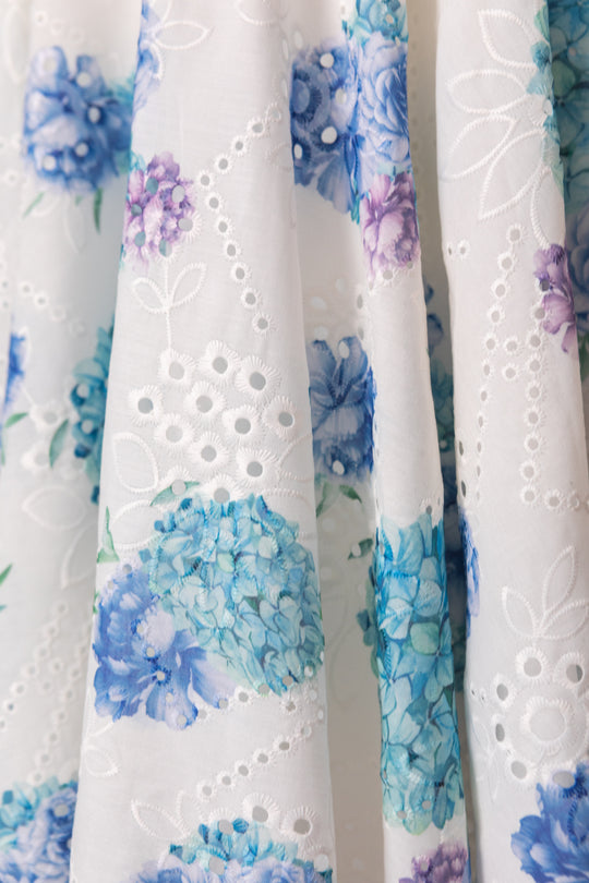 The Penny Dress in Blue Floral by Ivy City Co features white cotton eyelet fabric with blue and purple hydrangea prints, delicate cutouts, and stitched floral accents for a charming, feminine look.