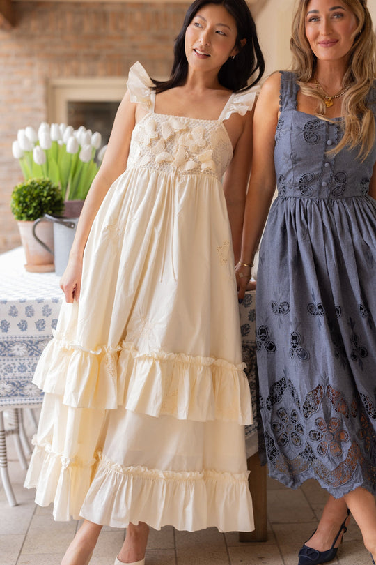 Two women stand indoors beside a table topped with a potted plant, posing for vibrant spring portraits. One wears the Gaia Butterfly Dress in Ivory by Ivy City Co—a cream ruffled maxi dress with embroidered details that is also maternity-friendly—while the other sports a blue sleeveless dress with floral cutouts. Both are smiling brightly.