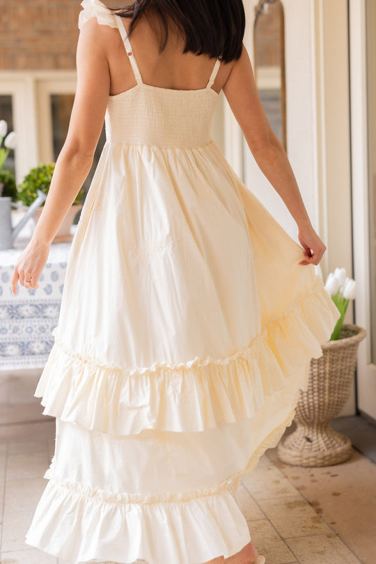 A woman with dark hair, wearing the Gaia Butterfly Dress in Ivory by Ivy City Co—a sleeveless, cream-colored ruffled maxi dress—stands indoors and lifts one side of her skirt. White tulips and a patterned tablecloth in the background create a perfect setting for spring portraits.