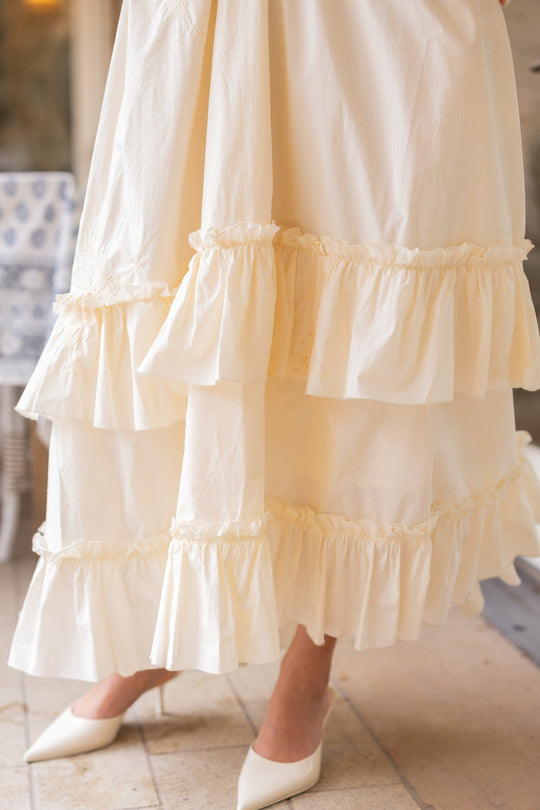 A close-up of the Ivy City Co Gaia Butterfly Dress in Ivory, featuring long, cream-colored ruffled layers and tiered details, brings a graceful touch perfect for spring portraits. Styled with pointed white heels and captured on an indoor tiled floor, this maternity-friendly dress offers both elegance and comfort.