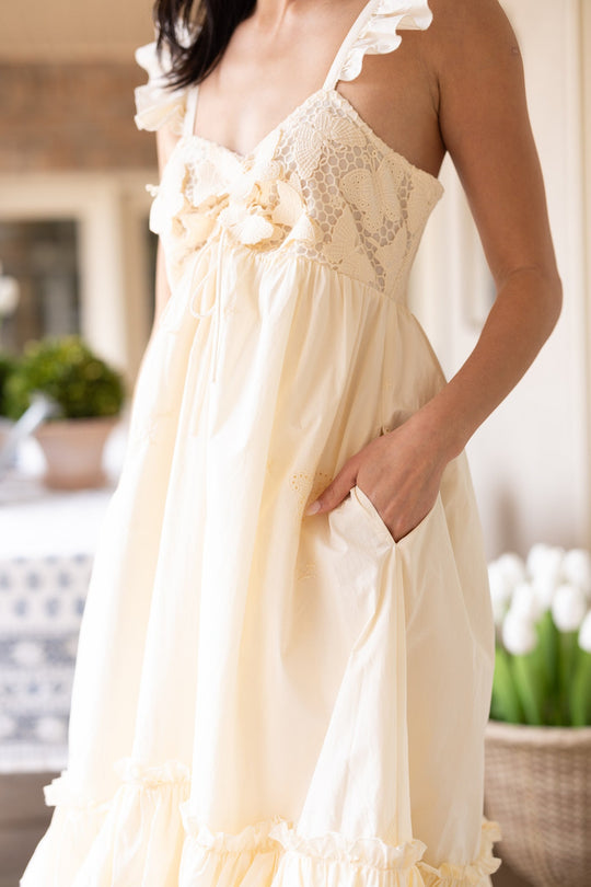 A woman wearing the Gaia Butterfly Dress in Ivory by Ivy City Co—a cream-colored, sleeveless, ruffled maxi dress with lace details and a pocket—stands indoors near a table adorned with plants and a basket of white tulips in the background. This look is perfect for spring portraits and beautifully maternity friendly.