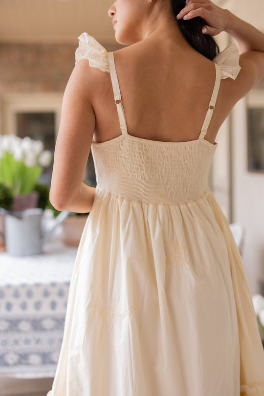 A woman stands indoors with her back to the camera, wearing the Gaia Butterfly Dress in Ivory by Ivy City Co. This cream-colored, sleeveless maxi dress features ruffled straps and gathered fabric—an ideal choice for spring portraits. She holds her dark hair in one hand, while a table and potted flowers appear in the background.