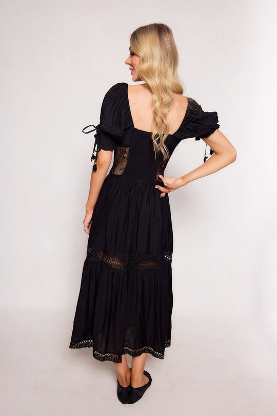 A woman with long blonde hair stands on a light background, wearing the Ivy City Co Peregrine Dress in Black—an ankle-length, short-sleeved dress featuring lace details and a low back—with one hand on her hip.