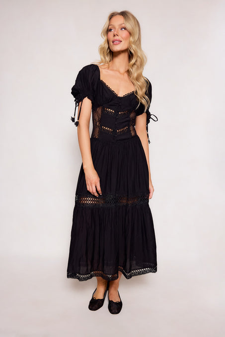 A woman with long blonde hair wears the Ivy City Co Peregrine Dress in Black, a lace-trimmed, puff-sleeve midi dress, paired with black flats. She stands against a plain light background, looking to the side and smiling.