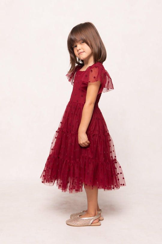 A young girl with brown hair stands in profile, wearing the Ivy City Co Mini Solange Dress in Burgundy Tulle. This girls’ holiday dress features layered textured polka dots, short sleeves, and a knee-length silhouette. She completes the look with beige sandals against a plain light background.