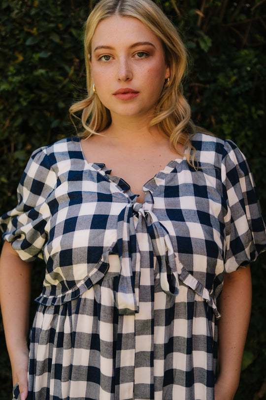 A blonde woman wears the Ivy City Co Picnic Dress in Navy Gingham with a tie neckline, standing before green foliage and looking directly at the camera with a neutral expression.
