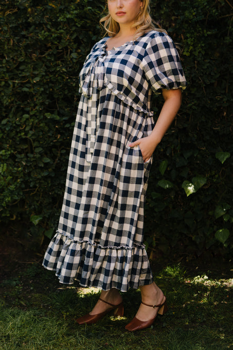 A person stands outdoors in the Ivy City Co Picnic Dress in Navy Gingham, featuring puff sleeves and a ruffled hem, paired with brown heeled shoes. This maternity-friendly look pops against the lush green grass and foliage—perfect for a picnic.