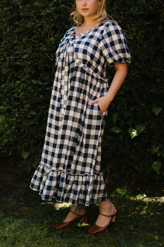 A person stands outdoors in the Ivy City Co Picnic Dress in Navy Gingham, featuring puff sleeves and a ruffled hem, paired with brown heeled shoes. This maternity-friendly look pops against the lush green grass and foliage—perfect for a picnic.