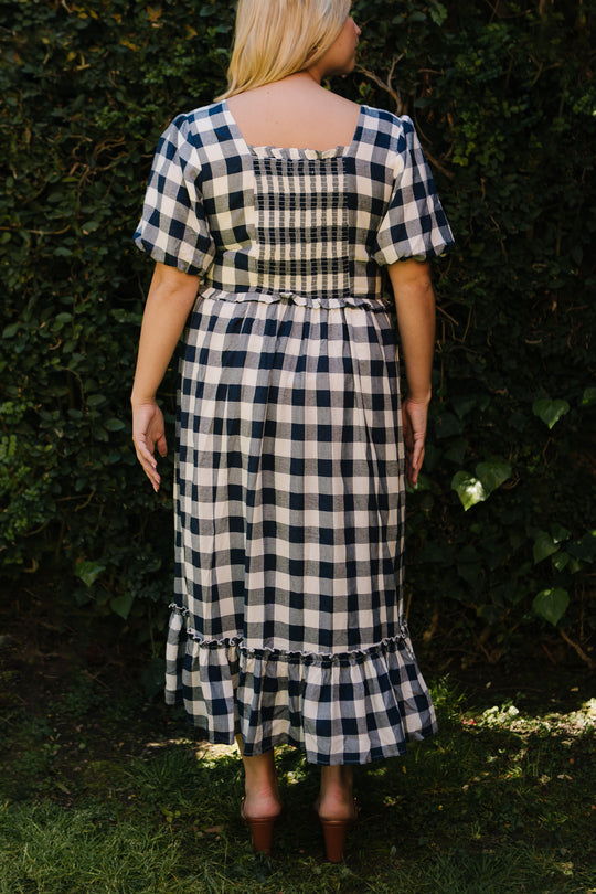 A woman stands outdoors with her back to the camera, wearing the Ivy City Co Picnic Dress in Navy Gingham. The dress features short sleeves, a smocked bodice, and a ruffled hem as she stands on grass before a leafy green backdrop.