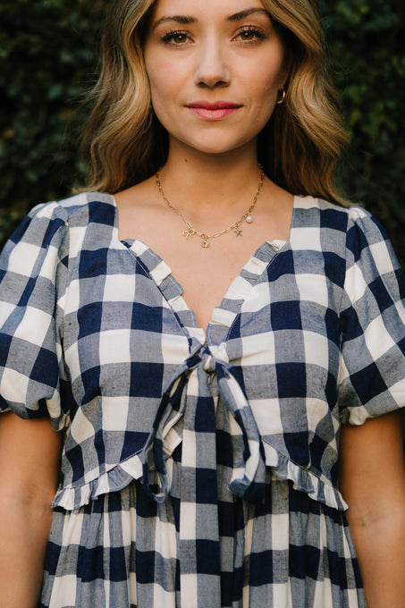 A woman with wavy blonde hair wears the Ivy City Co Picnic Dress in Navy Gingham, paired with a gold necklace. She stands in front of leafy greenery, smiling slightly and is photographed from the waist up.