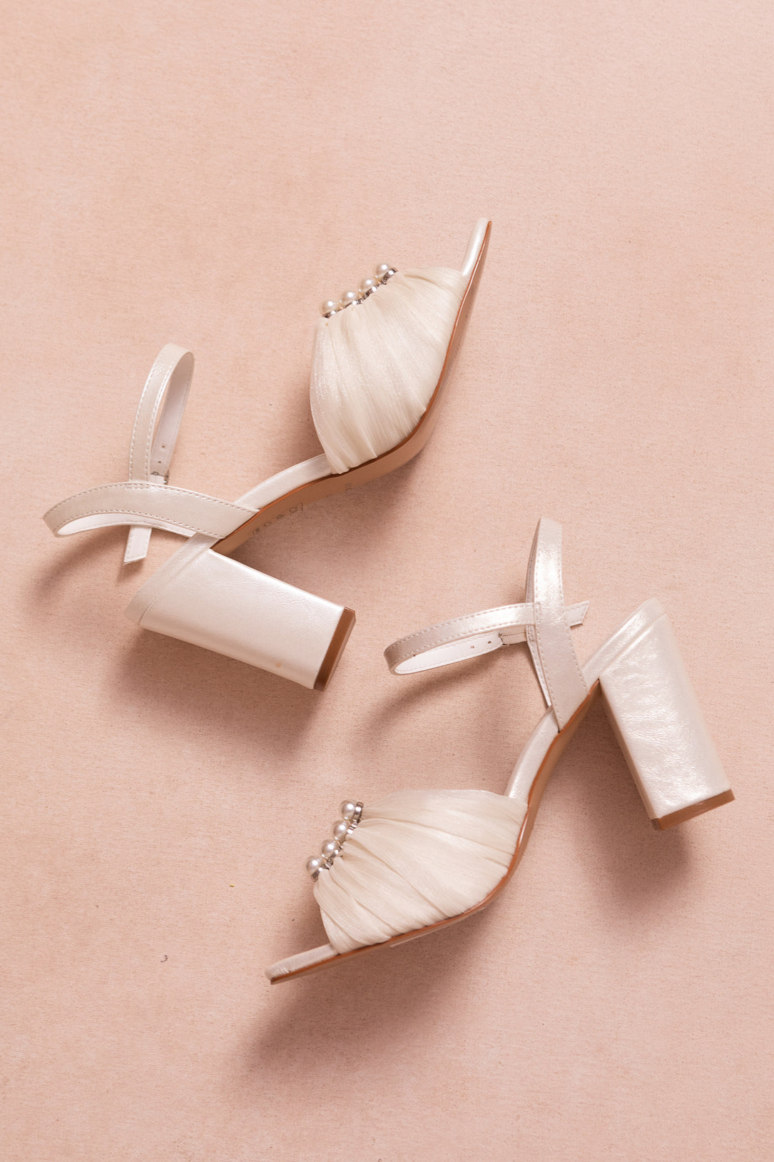 The Piper Pearl Heel in Ivory by Ivy City Co features elegant ankle straps, pleated front fabric, and pearl embellishments—perfect high-heeled sandals for any special occasion, shown against a soft beige background.