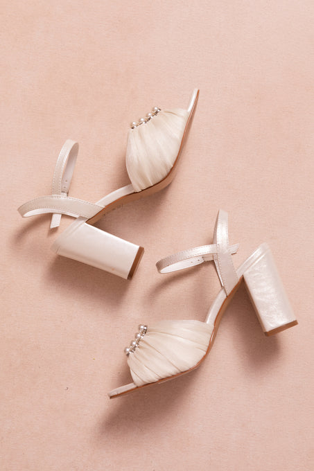 The Piper Pearl Heel in Ivory by Ivy City Co features elegant ankle straps, pleated front fabric, and pearl embellishments—perfect high-heeled sandals for any special occasion, shown against a soft beige background.