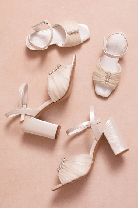 Three Ivy City Co Mini Piper Pearl Heel sandals in Ivory with block heels, ruched straps, and pearl accents are displayed on a light beige surface—one sandal is sideways and two face upward.