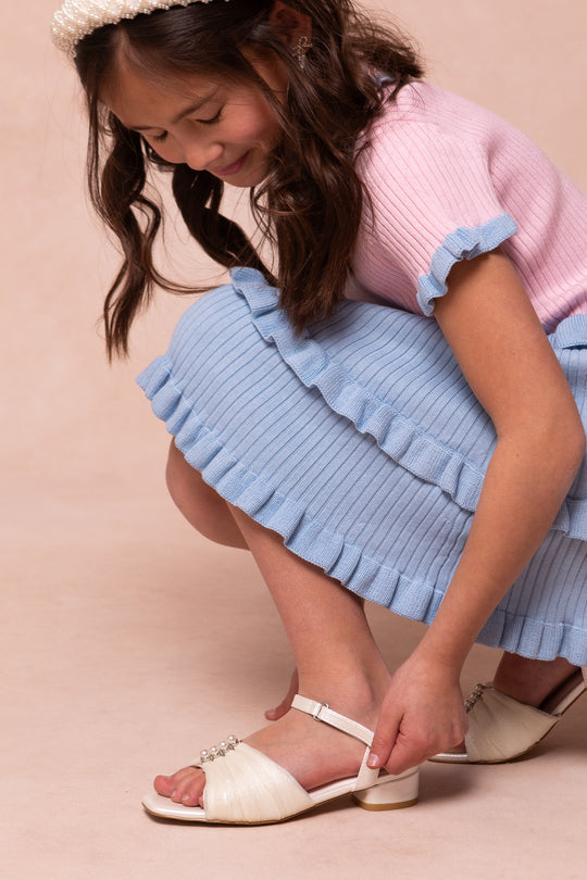 A young girl in a pink top and blue skirt smiles as she adjusts the strap on her Mini Piper Pearl Heel in Ivory by Ivy City Co—stylish vegan leather dress shoes with pearl details.